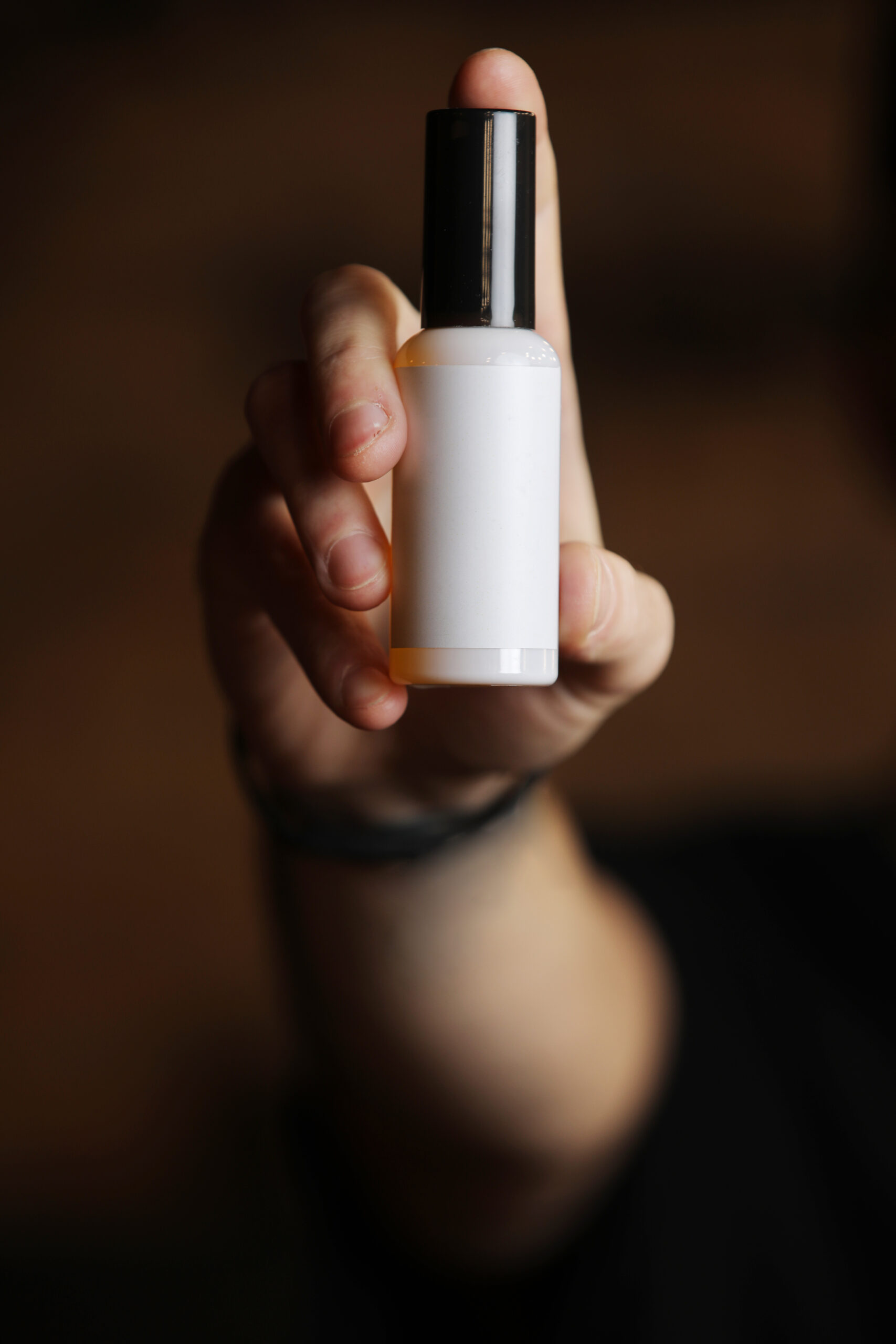 Unfocus young man holding in hand white plastic bottle with mock up for text on dark background. Focus on packaging bottle. Grooming for skin, hair men, beauty concept
